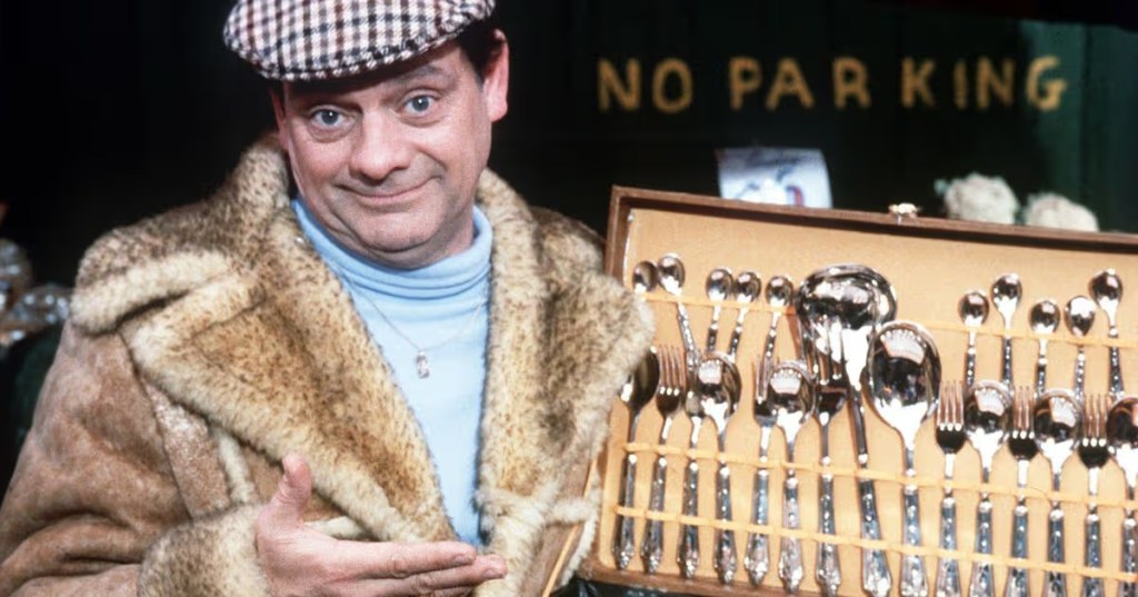 A man in a fur coat and a checkered hat smiling while showcasing a collection of silver cutlery in a wooden box, with a 'NO PARKING' sign in the background.
